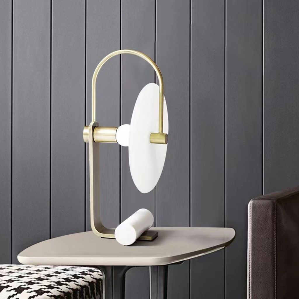 Đèn bàn chao thuỷ tinh với thiết kế tối giản mà sang trọng, sở hữu khung kim loại màu đồng ánh kim uốn cong mềm mại, kết hợp cùng chao đèn hình đĩa tròn mờ đục, tạo nên một bố cục cân bằng, hiện đại và đầy cá tính. Phần chân đèn thiết kế độc đáo với thanh trụ tròn bằng sứ trắng tinh khôi tạo điểm tựa vững chắc, vừa như một chi tiết trang trí tinh tế vừa đảm bảo độ ổn định cho sản phẩm.